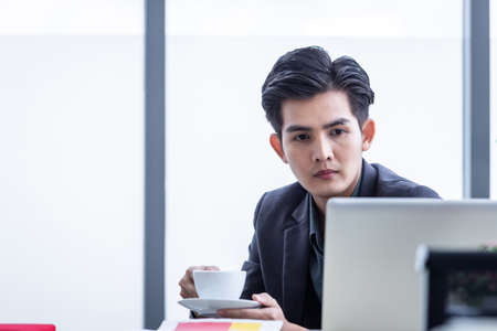 asian young businessman holding a coffee see a successful business plan on the laptop computer on...