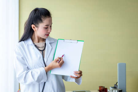 Asian young female doctor therapeutic advising with positive emotions hold empty clipboard and bed in hospital background,copy spaceの写真素材