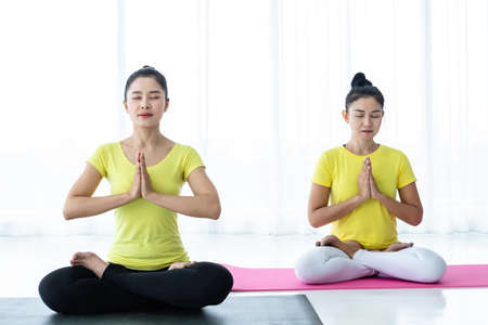 Two young Asian women workout practicing yoga in yellow dress or pose with a trainer and practice meditation wellness lifestyle and health fitness concept in a gym.の写真素材