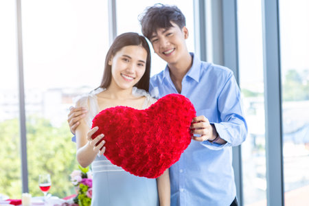Valentine's day concept,asian Young happy sweet couple in love holding a red heart shaped pillow After lunch In a restaurant background,Love story coupleの写真素材