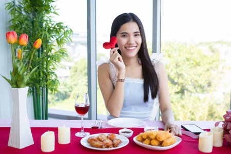 Valentine's day concept, Smiling Happy Asian young female sitting at a table food show holding small a red hearts shaped at in the restaurant backgroundの写真素材