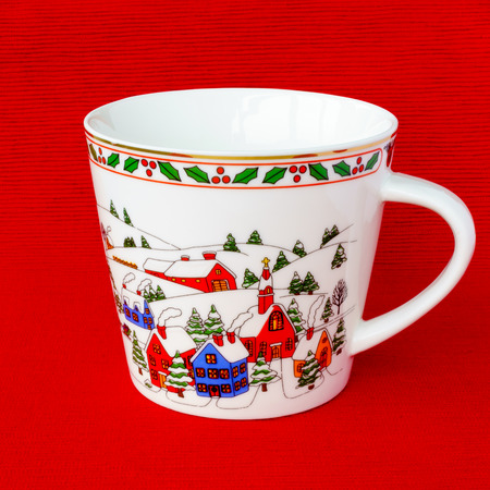 Brand new empty festive Christmas porcelain mug with colorful houses, green trees and a snowy landscape isolated on a red background.の写真素材