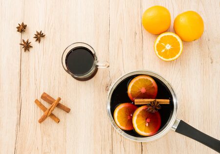 Stainless steel pot with hot, freshly prepared, homemade mulled wine, a drinking glass filled with mulled wine, cinnamon sticks, star anise and oranges on light brown wooden background.の写真素材