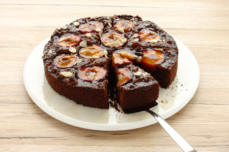 Closeup of a whole homemade Sticky Chocolate Plum Cake on a porcelain plate with one piece of cake on a cake server. Isolated on light brown wooden background.の写真素材