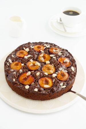 Closeup of a whole homemade Sticky Chocolate Plum Cake on a porcelain plate with a cake server on white background. In the background a cup of coffee, plate and milk jug. Shallow depth of field. の写真素材