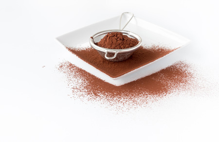 Close up of a metal sieve filled with cocoa powder on a white square porcelain plate. White background with copy space, shallow depth of field and selective focus on the cocoa powder in the sieve.の写真素材