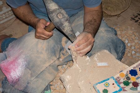 A moroccan craftsman is sitting on the floor making mosaics out of ceramic using a hammer.の写真素材