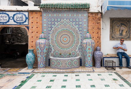 Fez, Morocco - May 07, 2017: Moroccan factory worker of the Mosaic Factory in Fez is sitting on a bench smoking a cigarette in the courtyard next to a beautiful mosaic fountain while having a break.のeditorial素材