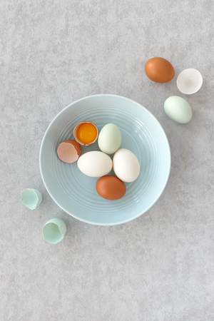 Blue eggs, brown eggs and white duck eggs, whole and halves, in a pastel blue bowl on grey textured...