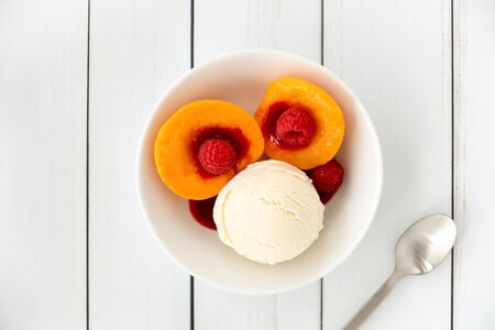 Top view of a Peach Melba sundae in a white porcelain bowl filled with vanilla ice cream, peaches, raspberries and raspberry sauce on white wood panel background with copy space.の写真素材