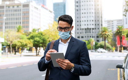 Young businessman wearing face mask and using digital tablet while walking in the streetの写真素材