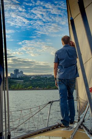 Ulyanovsk, Russia - July 20, 2019. The yachtsman is on the deck, holding the tackle, and looks at the city on the shore.のeditorial素材