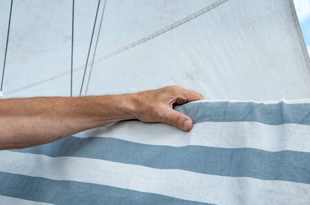 A male hand holding on to a yacht with a sheathed sail against the background of a deployed sail. A fragment of marine lifeの写真素材