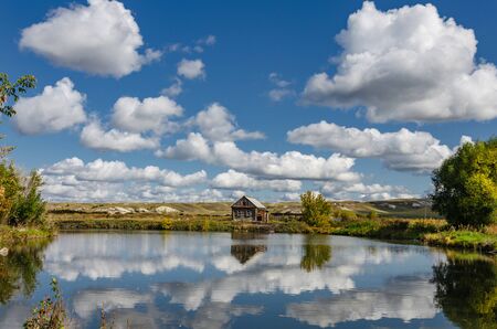 Photo of a lonely wooden house on the lake, which reflects summer clouds against the backdrop of...