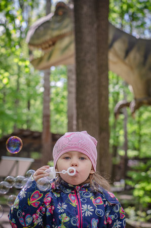 Ulyanovsk, Russia - may, 08. 2023: The girl enthusiastically blows soap bubbles while a dinosaur passes behind her behind the treesの写真素材