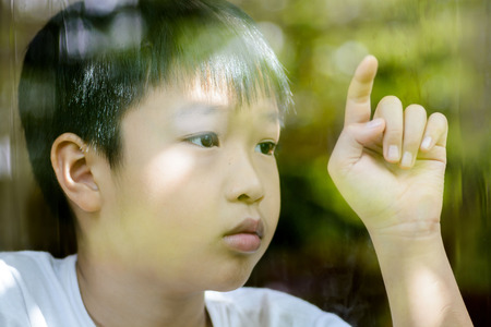 Vintage color tone, Lonely boy touch and write on glass window after rain look sad and wait for someone. Shooting from outside of glass window with tree reflect.の写真素材