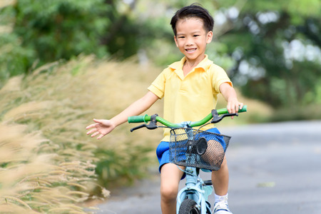 Young Asian boy ride a blue bicycle and touch the grass flower on the old road beside the tree and grass in summer time with warm sunlight.の写真素材