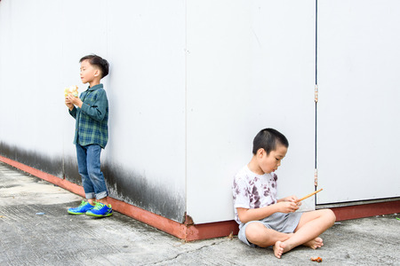 Younger Asian boy eat burger but poor boy eat chickent grill from bamboo stick at the corner of old building. Poor and hungry concept.の写真素材