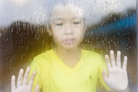 Young asian boy standing beside the window  with raindrop on the glass after raining with.の写真素材