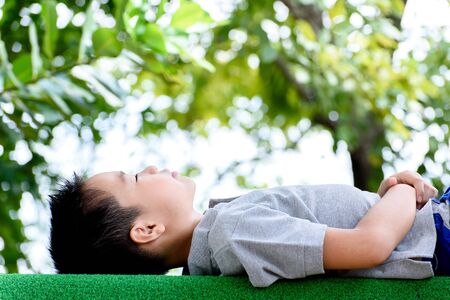 Young Asian boy lay down on the green lawn in the park during summer season with green bokehの写真素材
