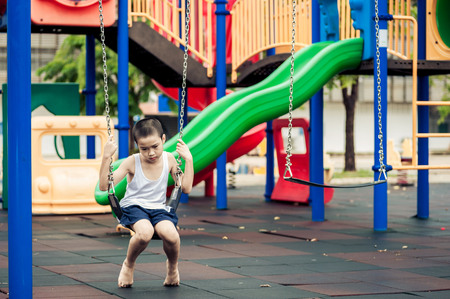 Dark color tone, unhappy young Asian boy sit and wating on swinging at outdoor playground.の写真素材