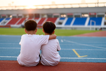 Two asian boy sit beside the blue running track and look to the stadium and sunset.の写真素材