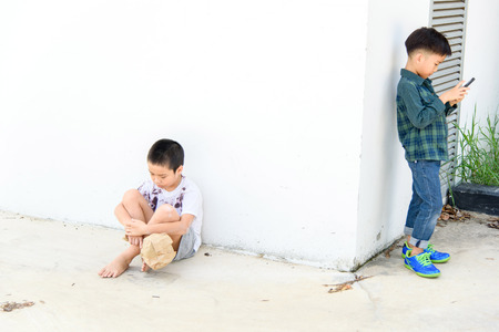 Younger Asian boy play smartphone but another poor boy looks unhappy and lonely at the white wall. Social network concept.の写真素材