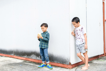 Younger Asian boy eat burger but poor boy eat chickent grill from bamboo stick at the corner of old building. Poor and hungry concept.の写真素材