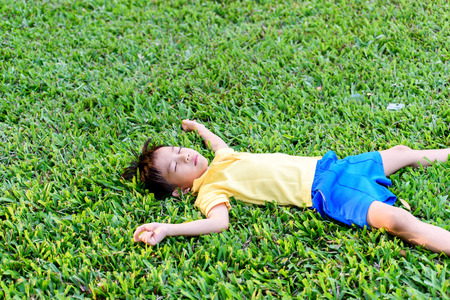 Young Asian boy in yellow shirt lay donw on green grass in the park during summer time.の写真素材
