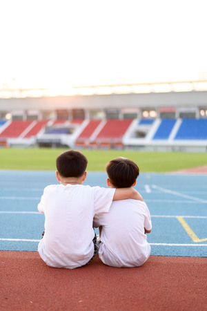 Two asian boy sit beside the blue running track and look to the stadium and sunset.の写真素材