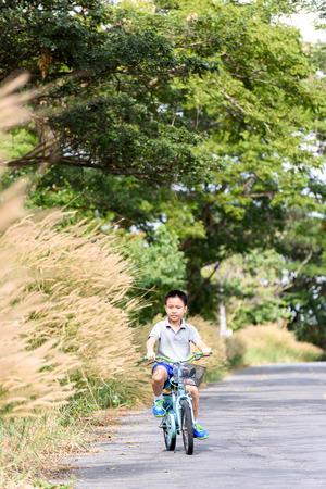 Young Asian boy ride a blue bicycle on the old road beside the tree and grass in summer time with warm sunlight.の写真素材