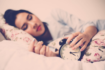 Vintage color tone. Selective focus on antique black alam clock in front of young and beautiful asian girl on the bed.の写真素材