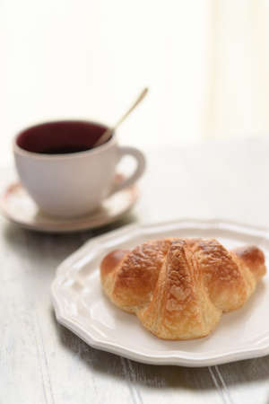 Selective focus on croissant brown skin. A fresh croissant on the white plate beside a cup of coffee on the wooden table.の写真素材