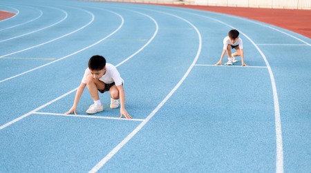 Young Asian boy prepare to start running with another boy on blue track in the stadium during day...