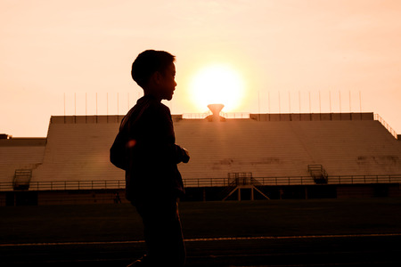 Silhouette Young boy in blue jacket running on track in the stadium against sunrise to practice himself.の写真素材
