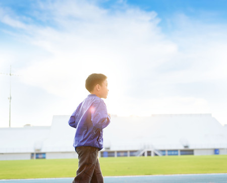 Young Asian boy in blue jacket running on track in the stadium against sunrise to practice himself.の写真素材