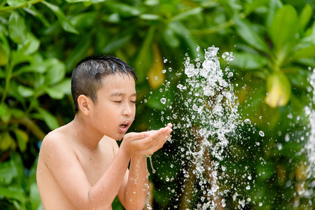 Young Asian Thai boy drink water from fountain tap with green tree background.の写真素材
