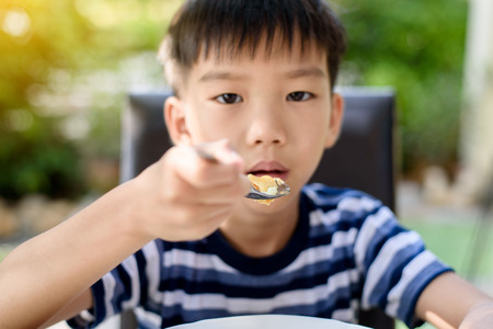 Selective focus at Young Thai boy eating with food on the wooden table with his family in the garden. And out focus tree background.の写真素材