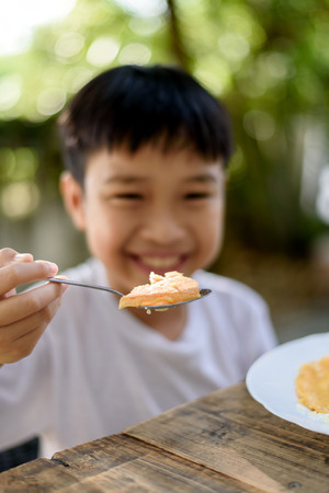 Selective focus at Young Thai boy eating with food on the wooden table with his family in the garden. And out focus tree background.の写真素材