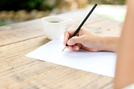 Selective close up at hand of woman writing on the white paper by pencil. Coffee cup beside on the wooden table/の写真素材