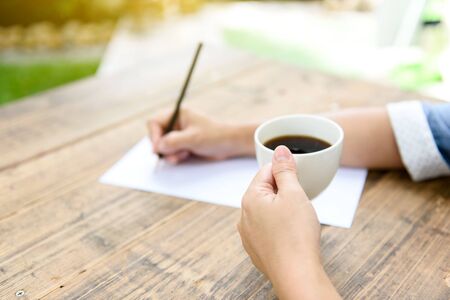 Selective close up at hand of woman writing on the white paper by pencil. Coffee cup beside on the wooden table/の写真素材