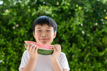 Young Asian boy eating watermelon in front of green garden background in the summer time with happy face.の写真素材