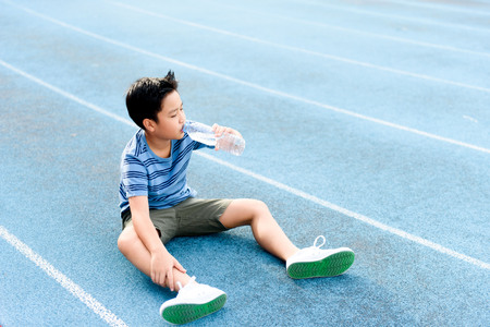 Young Asian Thai boy drinking water from bottle during resting on the blue track after runningの写真素材