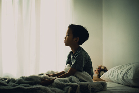 Dark tone color and sad mood of young Asian Thai boy sitting alone on a white bed in a white bedroomの写真素材