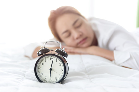 Selective focus at vintage black alarm clock morning time and out focus at young girl sleep on the white bed in bedroom.の写真素材