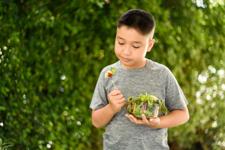 Selective focus of young Asian Thai boy eat salad in garden in front of gree tree background.の写真素材