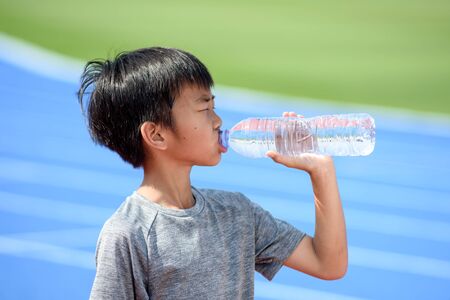 Young Asian boy drink water from a plastic bottle under the sunlight beside a running track to reduce his thirsty.の写真素材