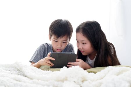 Young Asian boy and girl play a tablet phone on a bed.