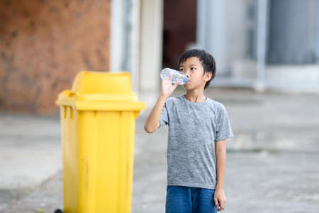 Young Asian boy drink water from plastic bottle in front of a yellow bin.の写真素材