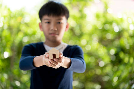 Selective focus at small wooden house in young boy hands in front of green background from tree in a...